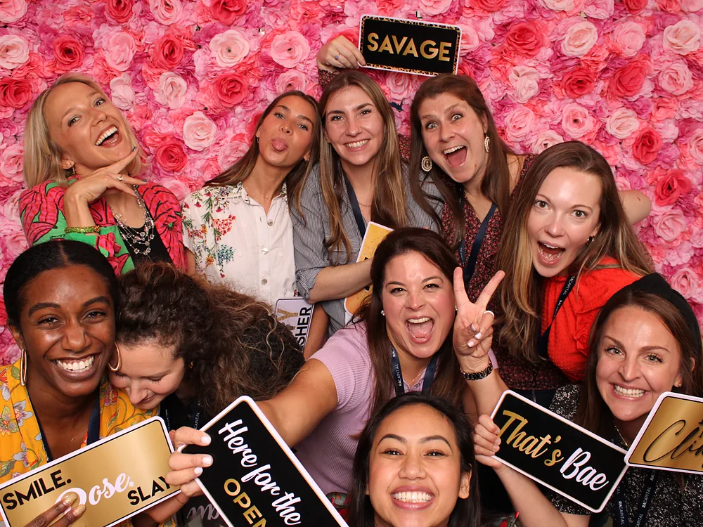 Large group of coworkers crowded together with prop signs in front of a pink flower wall