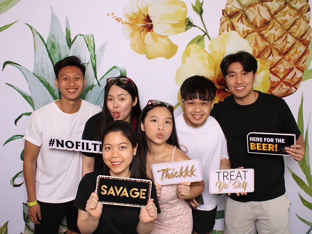 Friends holding prop signs in front of a tropical pineapple and hibiscus backdrop