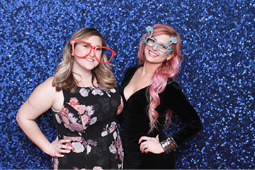 Two pretty females at a bar mitzvah party smile in front of a blue sparkle backdrop in the photo booth