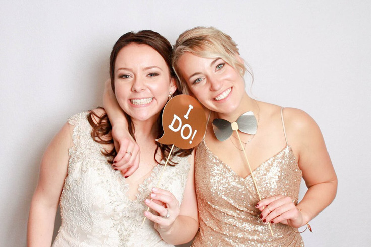 Bride Poses with Bridesmaid at photobooth with wedding themed props